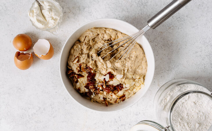 Banana cake batter being mixed in a bowl, with chopped dates and flaked almonds folded into the mixture.